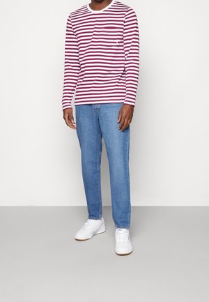 Mand iført en rød og hvid stribet langærmet skjorte, blå jeans og hvide sneakers, der står op ad en ensfarvet baggrund.