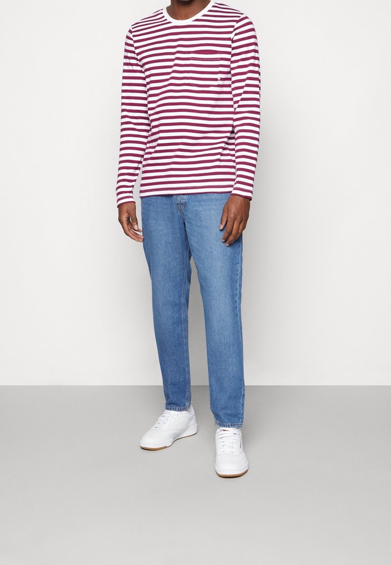Mann in einem rot-weißen gestreiften Langarmshirt, blauen Jeans und weißen Turnschuhen stehend vor einem einfarbigen Hintergrund.