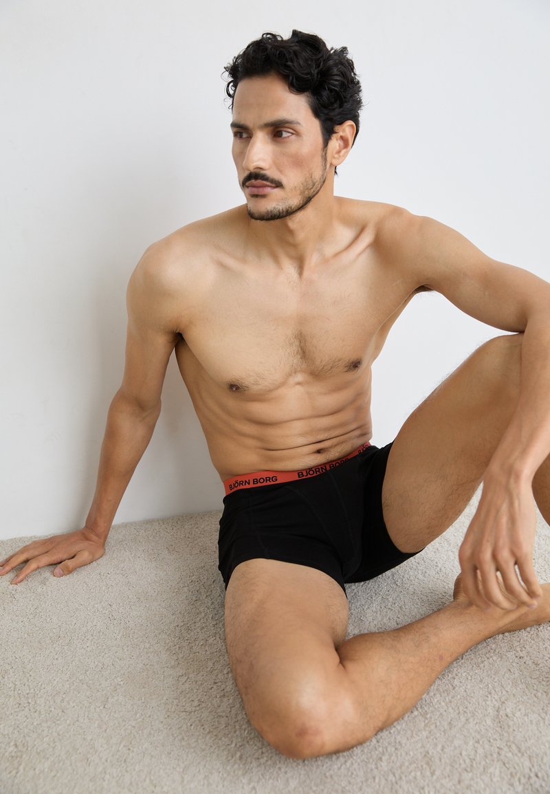 Fekete boxer alsónadrág piros derékpántos logóval, amelyet egy ülő férfi figura modellez egy világos szőnyegen, bemutatva az anyag textúráját és illeszkedését.