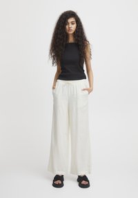 Zwarte geribbelde tanktop gecombineerd met brede crèmekleurige broek met een elastische tailleband en zakken, aangevuld met zwarte sandalen.