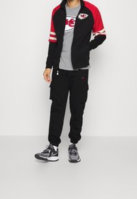 Mann in schwarzer und roter Kansas City Chiefs Jacke, grauem Logo-T-Shirt, schwarzen Cargo-Joggern und grauen Nike-Sneakern vor einfarbigem Hintergrund.