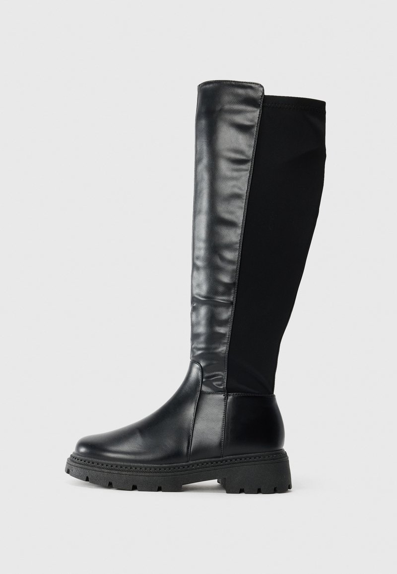 Botte noire montante jusqu'au genou avec devant en cuir lisse, dos en tissu extensible et semelle épaisse à crampons sur fond clair uni.