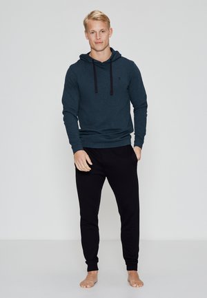 Mørk teal hættejakke med snoretræk, parret med sorte joggerbukser. Stoffet ser blødt ud, med en afslappet pasform og ankelsøm.