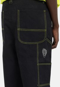 Black cargo pants made of sturdy fabric, featuring bright yellow stitching, multiple pockets, and a logo on the lower pocket.