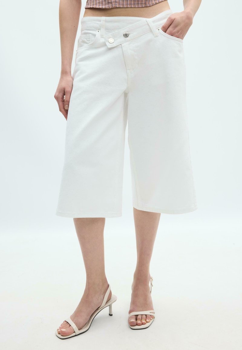 Femme portant un short blanc ample longueur genou avec fermeture asymétrique à boutons et sandales à talons beige à brides.
