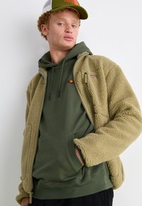 Chaqueta de forro polar marrón sobre una sudadera verde, con un logo y cordones ajustables. Una gorra verde con un logo completa el conjunto.