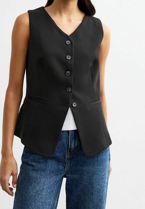 Kvinde iført en ærmeløs sort knapvest over en hvid top og blå jeans, vist fra skuldre til hofter mod en ensfarvet baggrund.