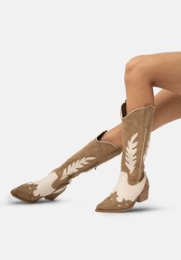 Kowbojskie buty ze skóry w odcieniach brązu i kremu, z szpiczastym noskiem, dekoracyjnymi wycięciami w kształcie liści oraz niskim, stosunkowo grubym obcasem. Zamek błyskawiczny po boku dla łatwego zakładania.