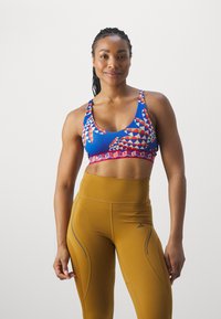 Sujetador con un patrón geométrico azul, acentos rojos y un ajuste ceñido, combinado con leggings mostaza de tiro alto que presentan detalles en la costura lateral.