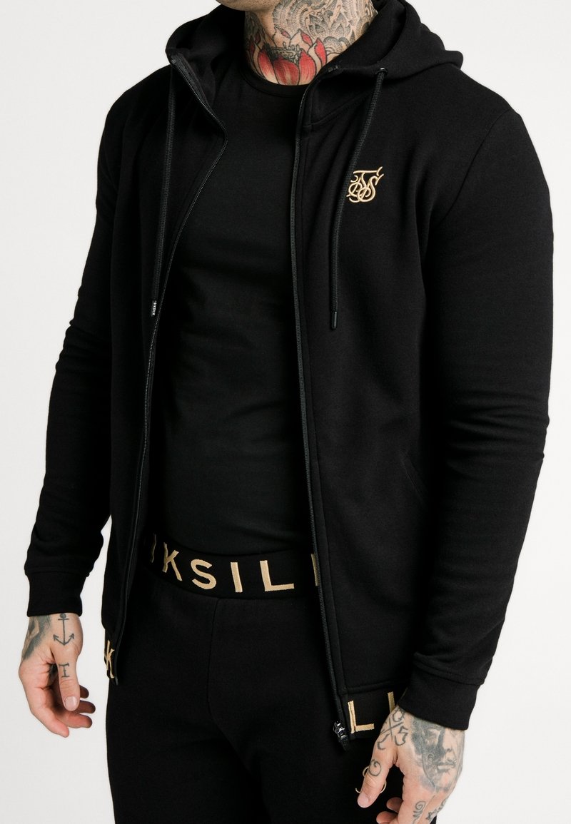 Homme portant un sweat à capuche noir zippé et un pantalon noir avec des inscriptions dorées, montrant des tatouages sur le cou et les mains sur fond blanc.