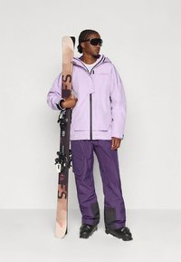 Lawendowa kurtka narciarska z czarnymi akcentami i kapturem, zestawiona z ciemnofioletowymi narciarskimi spodniami. Modelka trzyma snowboard z graficznym wzorem.