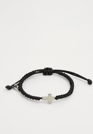 Mustassa punotussa rannekorussa on naru, johon on kiinnitetty metallinen hopeinen risti. Rannekorussa on säädettävä suljin, jossa on liukuva solmu.