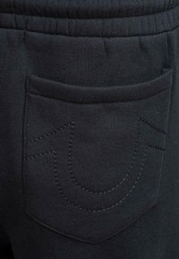 Tuvumā melna auduma bikses ar šūtu aizmugures kabatu, kurai ir izliekta divkārša šuvuma dizains, un elastīgu jostu.