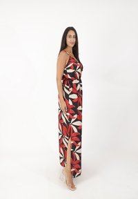 Robe maxi avec un motif floral noir, rouge et crème, sans manches, taille élastique et fente sur le côté ; associée à des sandales à talons transparents.