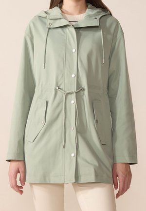 Mujer con impermeable verde claro con capucha, bolsillos delanteros, botones de presión y cintura ajustable con cordón, combinado con pantalones crema.