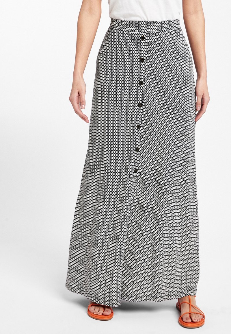 Femme portant une jupe longue à taille haute, à motifs noir et blanc avec des boutons sur le devant, et des sandales orange à lanières, debout devant un fond blanc.