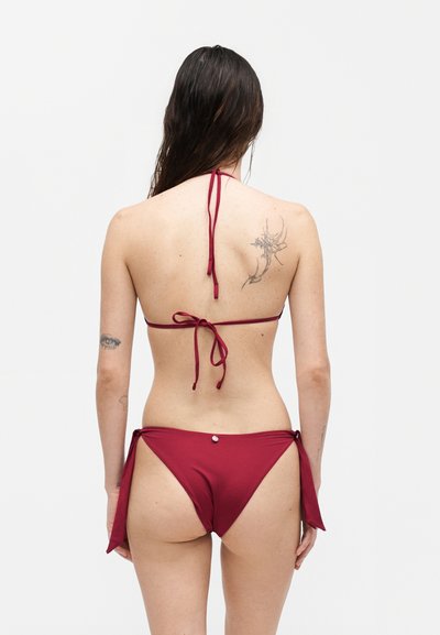 Γυναίκα με μακριά σκούρα μαλλιά που φοράει κόκκινο δεμένο μπικίνι, δείχνοντας την πλάτη με τατουάζ στον δεξί ώμο και αριστερό μπράτσο σε απλό φόντο.