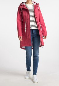 Kvinde iført en lang rød hættejakke med hvide snore, stribet skjorte, blå skinny jeans og hvide sneakers, stående foran en hvid baggrund.