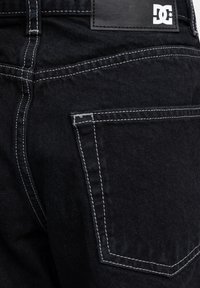 Pantalones de mezclilla negros con costuras blancas y un bolsillo trasero visible; cuentan con un parche de cuero texturizado con un logo en la cintura.