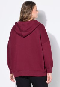 Sudadera oversized burdeos con capucha ajustable, hombros caídos y puños acanalados. Tejido suave y texturizado, corte casual.