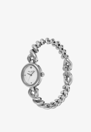 Zegarek w srebrnym odcieniu z masywną bransoletą w stylu łańcuszka oraz okrągłą białą tarczą. Posiada minimalistyczne znaczniki godzin oraz koronkę do ustawiania czasu.