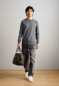 Pulover gri tricotat, blugi negri și adidași albi. Poartă o geantă verde măsliniu cu accesorii negre, stând pe un podea din lemn.