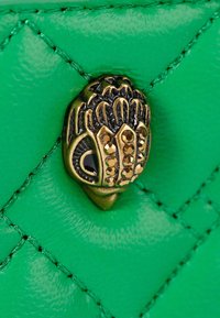Groen, gequilt leer met een gouden en zwarte decoratieve sluiting die verschillende vormen en ingelegde stenen heeft. Zachte textuur met contrasterende stiksels.