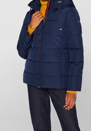 Osoba ubrana w granatową pikowaną kurtkę z kapturem, musztardowożółty golf i ciemnoniebieskie dżinsy, trzymająca rękę przy kołnierzu kurtki.