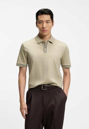 Giovane uomo con capelli neri corti che indossa una polo beige infilata in pantaloni plissettati marrone scuro con cintura nera.
