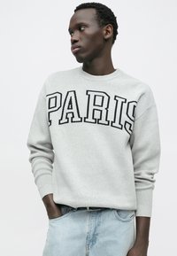 Pull gris clair avec "PARIS" en lettres noires, poignets côtelés et encolure ronde, associé à un jean en denim bleu. Texture lisse.