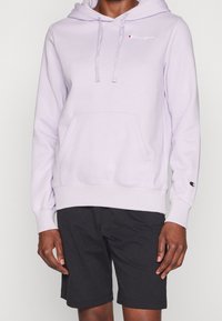 Person som bär en ljuslila Champion-hoodie med framficka och svarta shorts, synlig från nacken till midjan, mot en enfärgad bakgrund.