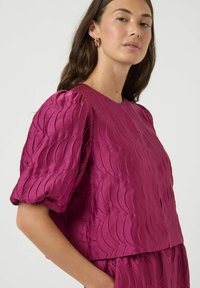 Fioletowa bluzka wykonana z teksturowanego satyny z falistymi wzorami. Bufiaste rękawy z marszczonymi detalami i okrągłym dekoltem.