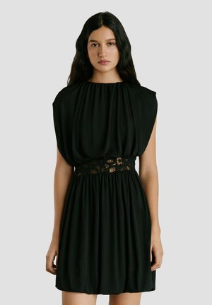 Vrouw met lang donker haar, gekleed in een mouwloze zwarte jurk met een geplooide lijfje en kanten details op de taille, staande tegen een effen achtergrond.