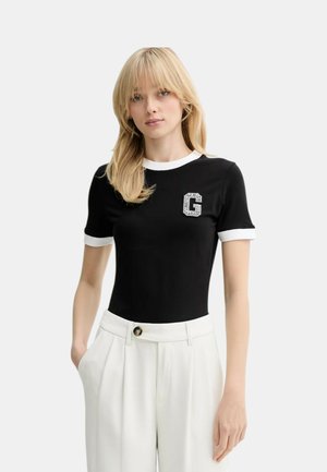 Donna con capelli biondi che indossa una maglietta nera a maniche corte con bordo bianco e una toppa con la lettera "G", abbinata a pantaloni bianchi a vita alta.