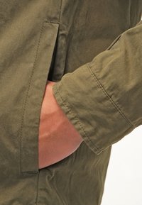 Olive green jacket with a smooth, matte fabric. Features a side pocket, long sleeves, and visible stitching details near the cuff.