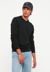 Hombre con suéter negro, jeans azul claro y gorra oscura, mirando hacia su izquierda contra un fondo blanco liso.