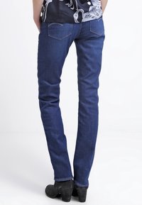 Úzké džíny z tmavě modrého denimu s jemným vyblednutím, zadními kapsami a minimálním prošíváním. Nošeny s černými kotníkovými botami.