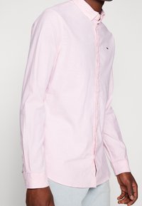 Mann trägt ein hellrosa langärmliges Hemd mit Knöpfen und einem kleinen Logo auf der Brust, kombiniert mit hellblauen Hosen.