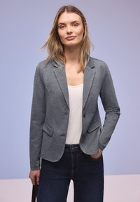 Jeune femme aux cheveux blonds détachés portant un blazer gris, un haut blanc et un jean foncé, debout devant un fond violet clair.