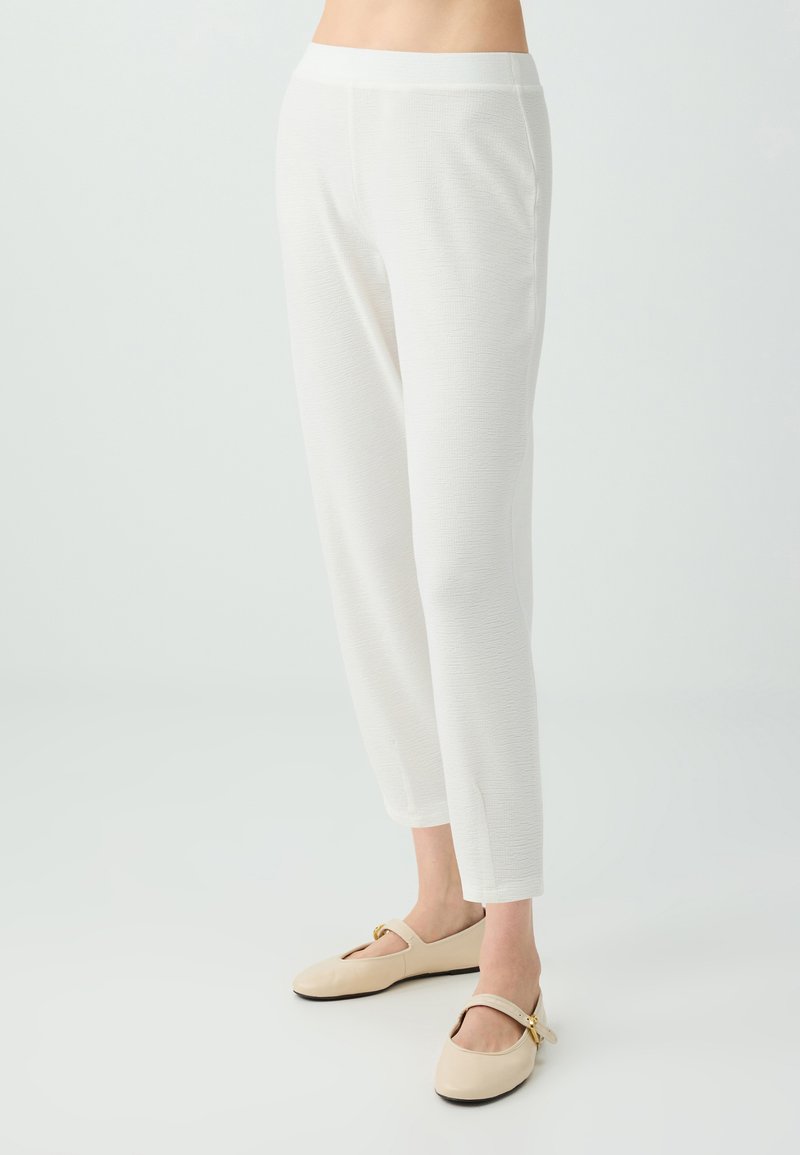 Femme portant un pantalon blanc texturé longueur cheville et des chaussures plates Mary Jane beiges, debout sur un fond clair uni.