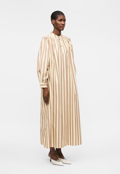 Γυναίκα που στέκεται φορώντας ένα μακρύ μπεζ και ανοιχτό καφέ φόρεμα με κάθετες ρίγες, φουσκωτά μακριά μανίκια και λευκές μυτερές γόβες.