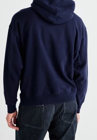 Felpa con cappuccio blu navy dal taglio comodo, con cuciture visibili e polsini a coste. Indossata con pantaloni scuri a righe.