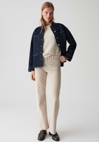 Dunkelblaue Jeansjacke mit Knopfverschluss, cremefarbener, strukturierter Pullover, beige Schlaghose und dunkelbraune Slippers. Schlichtes, modernes Ensemble.