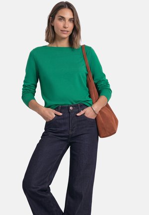 Femme portant une chemise verte à manches longues et un jean bleu foncé, mains dans les poches et un sac à bandoulière marron sur fond blanc.