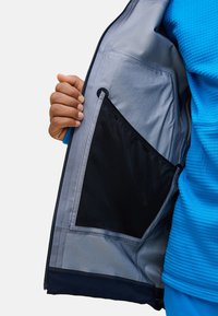 Blaue Jacke mit grauem Innenfutter, ausgestattet mit einer großen schwarzen Innentasche mit elastischem Rand. Hand greift hinein und zeigt die Details der Tasche.