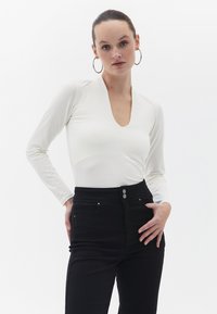 Langærmet hvid bodysuit med dyb V-hals, parret med højtaljede sorte jeans med to sølvfarvede knapdetaljer.