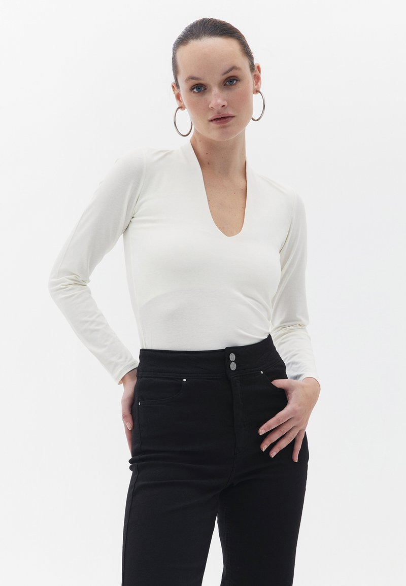 Langærmet hvid bodysuit med dyb V-hals, parret med højtaljede sorte jeans med to sølvfarvede knapdetaljer.