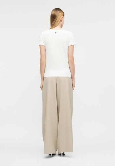 Γυναίκα με μακριά ξανθά μαλλιά που φοράει εφαρμοστό λευκό τοπ με κοντά μανίκια και μπεζ παντελόνι με φαρδιά πόδια, στέκεται γυρισμένη με την πλάτη σε λευκό φόντο.