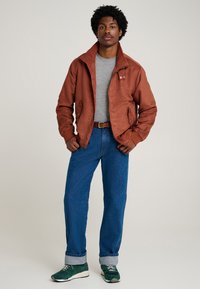 Jachetă bomber de culoarea ruginii, cu guler înalt, tricou gri, blugi albaștri și adidași verzi. Jacheta are două buzunare frontale.