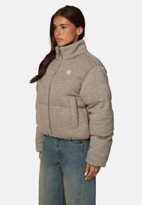 Kurtka puchowa w jasnobrązowym, teksturowanym materiale z wysokim kołnierzem, przednim zamkiem błyskawicznym i bocznymi kieszeniami. Posiada małe logo na piersi.